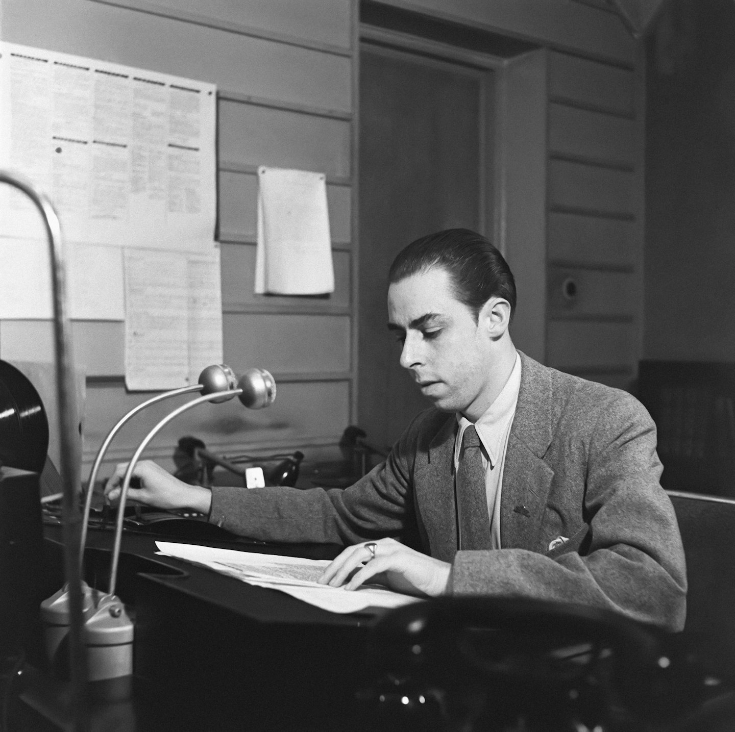Man in suit reviews papers at a desk with vintage lamp.